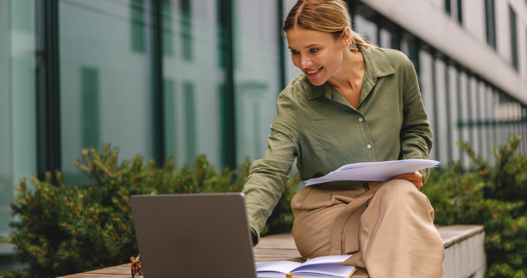 A woman sitting outside an office building works on her computer.
